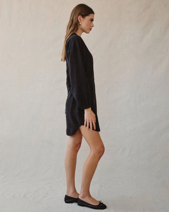 A woman stands in profile against a neutral background wearing the Bella Dahl Long Sleeve Belted Mini Dress in Black with black flats. Her hair is down and she faces left.