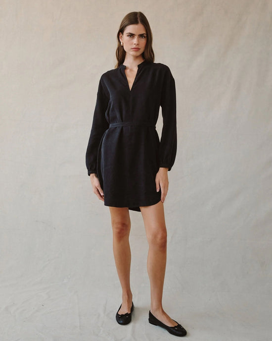 A woman with straight brown hair and a neutral expression stands against a plain beige background, wearing the Bella Dahl Long Sleeve Belted Mini Dress in Black and black flats, exuding effortless elegance.