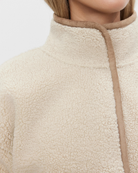 Close-up of a person wearing a high-collared, cream-colored, faux-shearling Albany Mock Neck Jacket in Sand with brown trim by Velvet By Graham & Spencer.