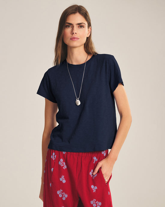 A woman with long brown hair wears the Marika Short Sleeve Crew Neck Crop Top in Midnight by Velvet by Graham & Spencer, a silver pendant necklace, and red pants with a small blue floral pattern, standing against a plain light background.