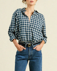 A woman wears the Trovata Birds of Paradis Blake Oversized Shirt in Maritime Plaid with rolled sleeves, tucked into high-waisted blue jeans and a black belt, standing against a plain background.