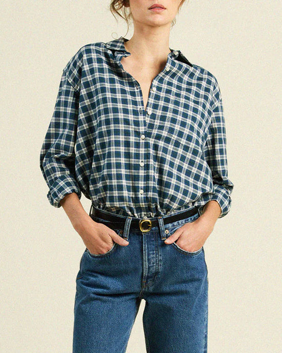 A woman wears the Trovata Birds of Paradis Blake Oversized Shirt in Maritime Plaid with rolled sleeves, tucked into high-waisted blue jeans and a black belt, standing against a plain background.