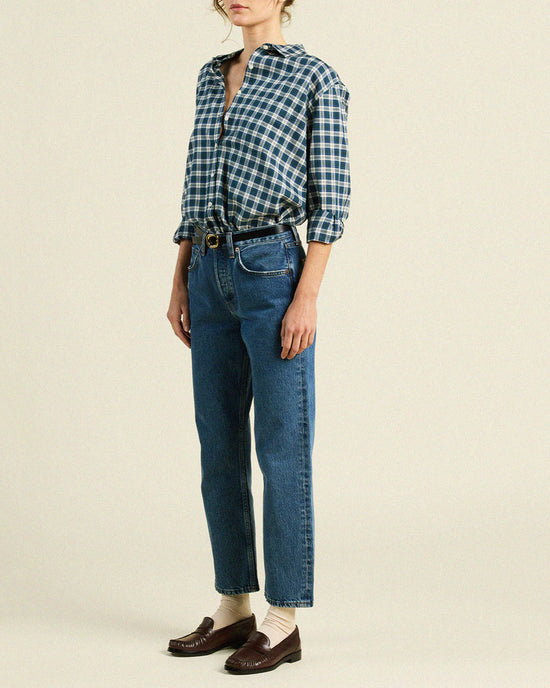 Someone wears the Trovata Birds of Paradis Blake Oversized Shirt in Maritime Plaid, tucked into high-waisted blue jeans with a black belt, beige socks, and brown loafers, standing against a plain background.