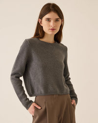 A woman with long brown hair wears the Demylee Melina Cotton Cashmere Sweater in Dark Heather Grey and brown trousers, standing against a plain light background with her hands in her pockets, looking directly at the camera.