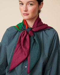 The person is styled in a green shirt, elegantly complemented by an oversized bandana, specifically the Bandana No 751 in Poppy Red by Moismont. This large, colorful scarf tied at the neck features a luxurious blend of wool, silk, and cashmere in vibrant shades of red, green, and orange.