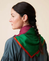 A woman with dark braided hair and gold hoop earrings, adorned in a green, red, and orange oversized bandana that captures the elegance of Moismont's Bandana No 751 in Poppy Red, gazes to the left. She is clothed in a dark blouse set against a neutral backdrop.