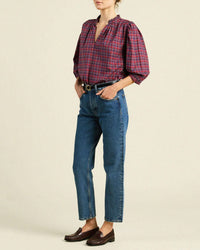 Wearing the Orly Blouse in Mulled Wine Plaid by Trovata Birds of Paradis, paired with high-waisted blue jeans, a black belt, and brown loafers, hands in pockets against a plain background.