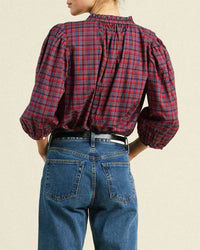 A person wearing the Trovata Birds of Paradis Orly Blouse in Mulled Wine Plaid with puffed sleeves paired with blue denim jeans, seen from behind. The blouse is tucked in and cinched with a black belt.