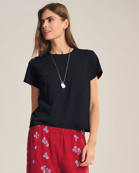 A woman with light brown hair wears the Velvet by Graham & Spencer Marika Short Sleeve Crew Neck Crop Top in black, paired with red floral pants and a pendant necklace, standing against a plain background.