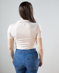 A woman with long brown hair stands facing away from the camera, wearing the NSF Alden S/S Fitted Snap Polo in Pearl and high-waisted blue jeans against a plain light background.
