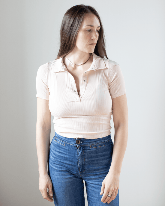 A woman with long brown hair wears the NSF Alden S/S Fitted Snap Polo in Pearl and blue jeans, standing against a plain white background. She looks slightly to her left with a neutral expression.