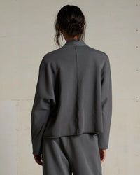 A person with long brown hair tied back, wearing the perfectwhitetee Olivia Fleece Short Cardigan in Charcoal and matching pants, stands facing away from the camera against a plain light-colored wall.