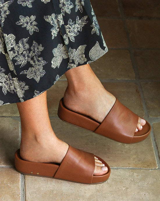 A person wearing a black skirt with white floral patterns and chic beek.. Pelican in Tan slides stands on tan tiled flooring.
