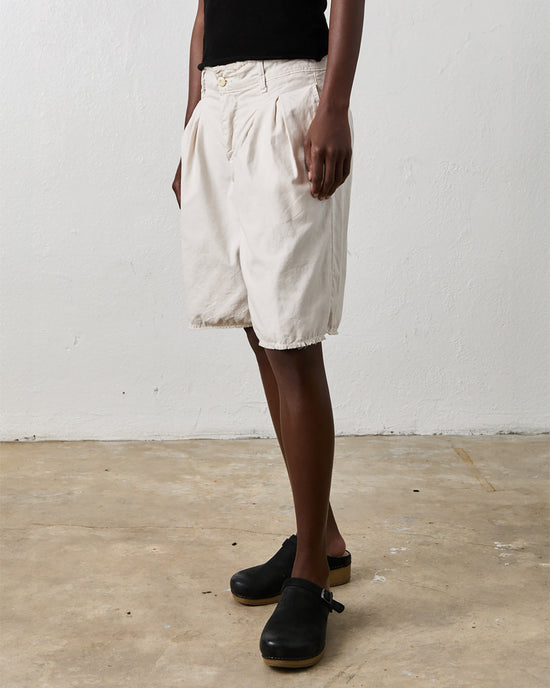 Person wearing white, Quinn Culotte in Soft White by NSF and black shoes stands against a plain white wall and concrete floor. The person is also dressed in a black top, embodying an effortlessly urban chic vibe.