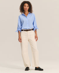 A woman with curly hair wears the Arlette Knot Sleeve Button Up in Chill by Velvet by Graham & Spencer, paired with cream wide-leg pants, a black belt, and black loafers, standing against a plain light background.