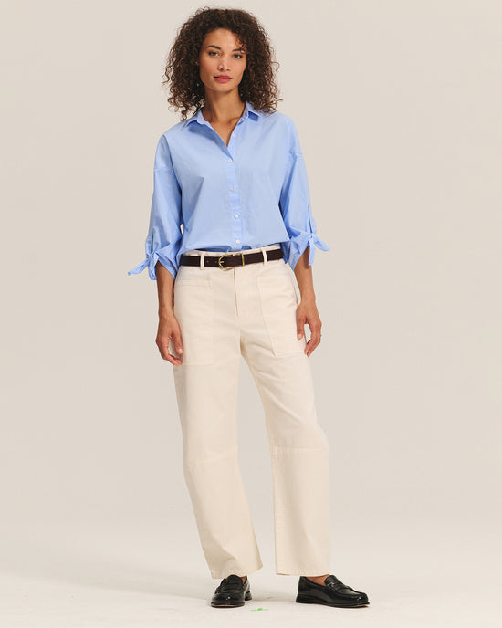 A woman with curly hair wears the Arlette Knot Sleeve Button Up in Chill by Velvet by Graham & Spencer, paired with cream wide-leg pants, a black belt, and black loafers, standing against a plain light background.