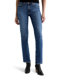 A person wearing AG Jeans’ Sandra Low Rise Straight in 15Ys Prague, a black top, and black heeled ankle boots stands against a white background.