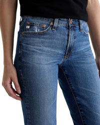 A person wears the Sandra Low Rise Straight in 15Ys Prague by AG Jeans, shown from waist to mid-thigh. The medium blue jeans feature visible stitching, front pockets, and copper rivets for a classic look.