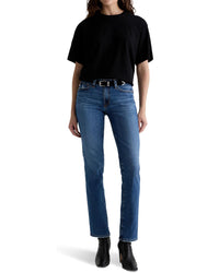 A person wearing a black t-shirt, Sandra Low Rise Straight in 15Ys Prague jeans by AG Jeans with a black belt, and black ankle boots, standing against a plain white background.