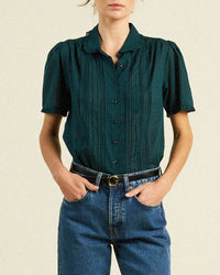 A person wears a Trovata Birds of Paradis Millie Blouse in Sea Pine Check with puffed shoulders and blue high-waisted jeans, hands in pockets, standing against a plain light background.