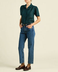 A person stands with hands in pockets, wearing the Trovata Birds of Paradis Millie Blouse in Sea Pine Check, blue high-waisted jeans, a black belt, and brown loafers against a plain light background.