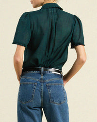 A person stands facing away, wearing a Trovata Birds of Paradis Millie Blouse in Sea Pine Check and high-waisted blue jeans with a black belt, hands on hips against a plain light background.