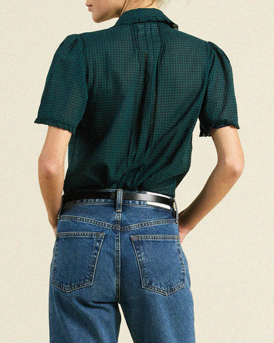 A person stands facing away, wearing a Trovata Birds of Paradis Millie Blouse in Sea Pine Check and high-waisted blue jeans with a black belt, hands on hips against a plain light background.
