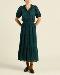 A person in the Blair Dress in Sea Pine Check by Trovata Birds of Paradis—short sleeves, fitted waist, tiered midi skirt—wears brown loafers and mustard yellow socks, standing against a plain light background.