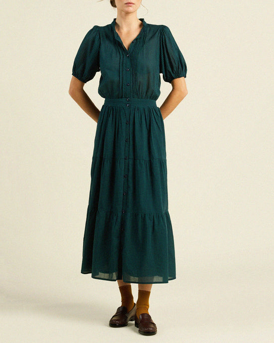 A person in the Blair Dress in Sea Pine Check by Trovata Birds of Paradis—short sleeves, fitted waist, tiered midi skirt—wears brown loafers and mustard yellow socks, standing against a plain light background.