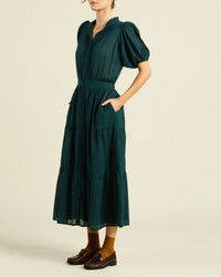 A woman wears the Trovata Birds of Paradis Blair Dress in Sea Pine Check with short puff sleeves, button-down front, pockets, and a tiered midi skirt. She pairs it with brown socks and loafers, standing against a plain light background. Her head is slightly out of frame.