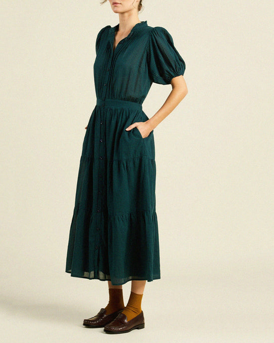 A woman wears the Trovata Birds of Paradis Blair Dress in Sea Pine Check with short puff sleeves, button-down front, pockets, and a tiered midi skirt. She pairs it with brown socks and loafers, standing against a plain light background. Her head is slightly out of frame.