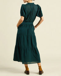A person stands facing away, wearing the Blair Dress in Sea Pine Check by Trovata Birds of Paradis, featuring puffed short sleeves and a tiered midi skirt. One hand is in a pocket and they pair it with brown shoes and mustard-yellow socks.