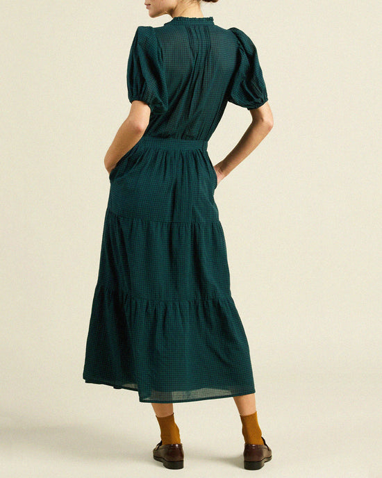A person stands facing away, wearing the Blair Dress in Sea Pine Check by Trovata Birds of Paradis, featuring puffed short sleeves and a tiered midi skirt. One hand is in a pocket and they pair it with brown shoes and mustard-yellow socks.