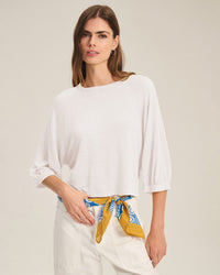 A woman with long brown hair stands against a plain light background, wearing the Shelly Long Sleeve Top in White by Velvet by Graham & Spencer, paired with white pants and accessorized with a yellow and blue patterned scarf at her waist.