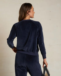 A woman stands against a light wall, holding a bag in her left hand and wearing the Stella Velour Shrunken Raglan SS sweatshirt in Navy by perfectwhitetee, paired with matching pants for a luxe, relaxed look.