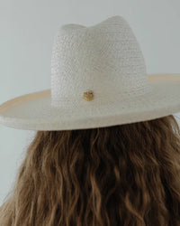 A person with long, wavy brown hair wearing a wide-brimmed, textured Gigi Pip Cara Loren Pencil Brim Straw hat in White with a small golden emblem at the front.