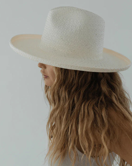 Profile of a woman with long wavy hair, wearing a wide-brimmed Gigi Pip Cara Loren Pencil Brim Straw in White hat, against a light gray background.
