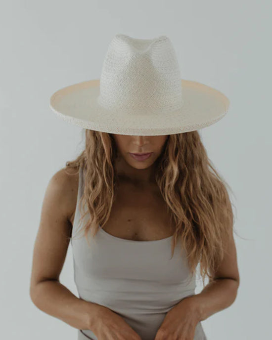 A woman in a Cara Loren Pencil Brim Straw in White by Gigi Pip, wearing an adjustable straw hat that covers her eyes, against a plain background.