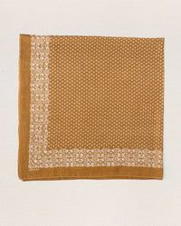 The Big Bandana in Marigold by the Great is a square brown cotton bandana, inspired by vintage handkerchiefs, with a white floral and dot border and small white center dots, displayed on a textured light surface.