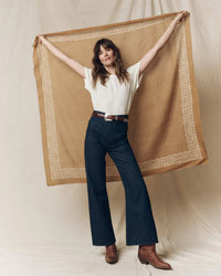 A woman smiles, holding The Big Bandana in Marigold by the Great behind her. She wears a white t-shirt, dark wide-leg jeans, a belt, and brown boots against a neutral fabric backdrop.