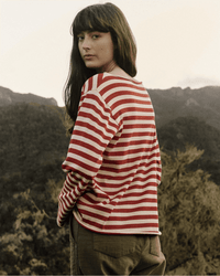 A woman with long dark hair wears the Great’s Sailor Sweater in Maple Stripe and brown pants, standing outdoors with her back mostly turned, looking over her shoulder against a backdrop of mountains and trees.
