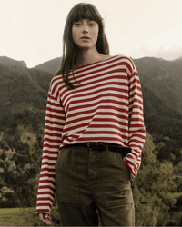 A woman with long brown hair stands outdoors with mountains and trees behind her, wearing the Great's The Sailor Sweater in Maple Stripe and olive green pants.