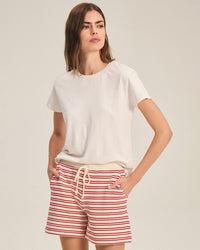A woman stands with her hands in her pockets, wearing Velvet by Graham & Spencer's Tia Short Sleeve Crew Neck Top in Beach and red-and-white striped drawstring shorts, looking off to the side against a neutral background.