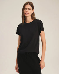 A woman with long brown hair wears the Velvet by Graham & Spencer Tia Short Sleeve Crew Neck Top in Black and a black skirt, standing against a neutral beige background.
