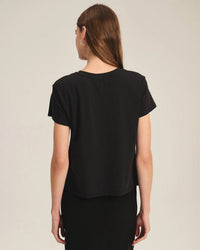 A woman with long brown hair stands with her back to the camera against a plain light background, wearing the Velvet by Graham & Spencer Tia Short Sleeve Crew Neck Top in Black and a black skirt.