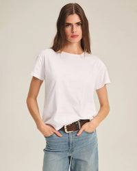 A woman with long brown hair wears the Tia Short Sleeve Crew Neck Top in White by Velvet by Graham & Spencer, paired with blue jeans and a brown belt, standing against a neutral background with her hands in her pockets.