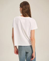 A woman with long brown hair faces away, wearing the Velvet by Graham & Spencer Tia Short Sleeve Crew Neck Top in White and light blue jeans, set against a neutral background.