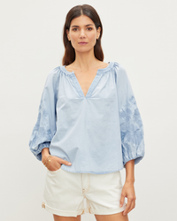Woman in a Velvet by Graham & Spencer Trina L/S Boho Top in Chambray and white shorts posing against a plain background.