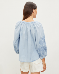 Woman wearing a Velvet by Graham & Spencer Trina L/S Boho Top in Chambray with floral embroidered details and white shorts, standing with her back to the camera.