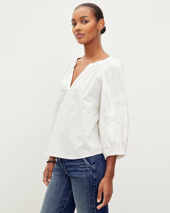 A woman posing in a Trina L/S Boho Top in Off White by Velvet by Graham & Spencer and blue jeans against a neutral background.